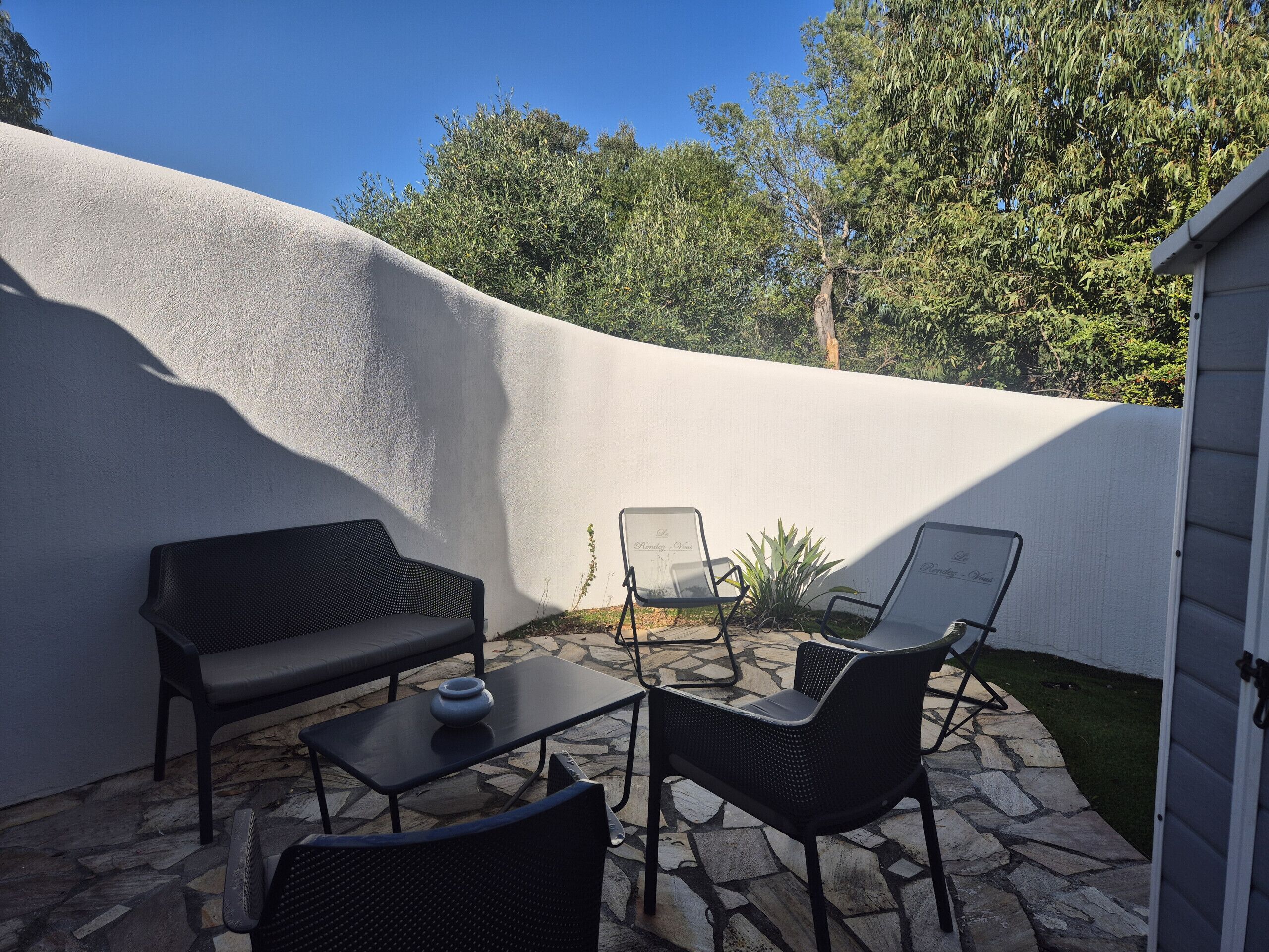 Eine Terrasse mit Steinboden, einem schwarzen Sofa, zwei schwarzen Stühlen und einem kleinen Tisch.