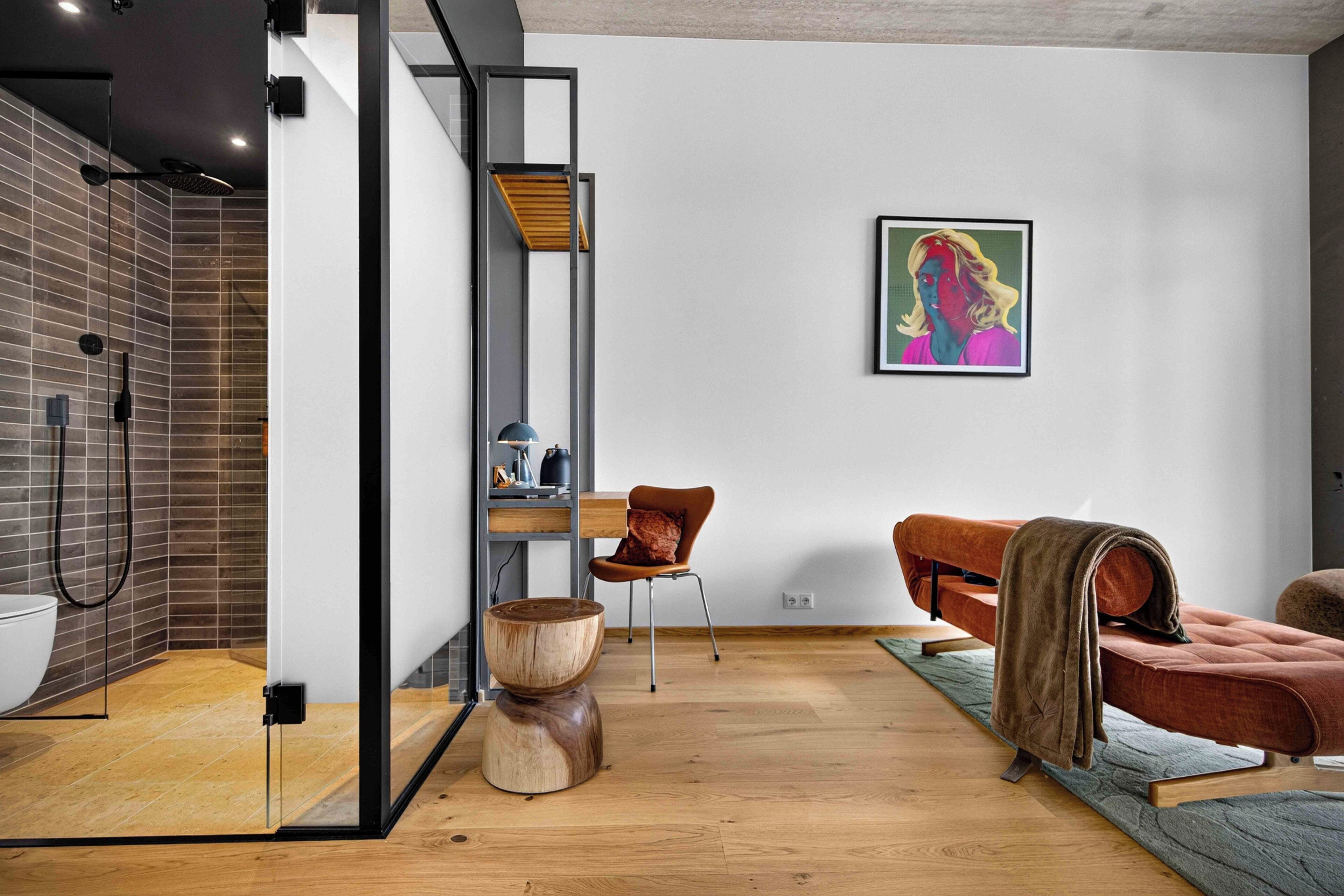 A modern interior with a wooden floor, a glass partition, a brown leather sofa, a wooden coffee table, and a colorful painting on the wall.