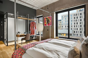 A modern hotel room with a bed, a desk, a chair, a wardrobe, and a large window with a city view.