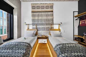 Una habitación de hotel con un gran tapiz tejido en la pared, dos camas con ropa de cama estampada a juego, una mesita de noche de madera, una lámpara de noche y un perchero con una maleta.