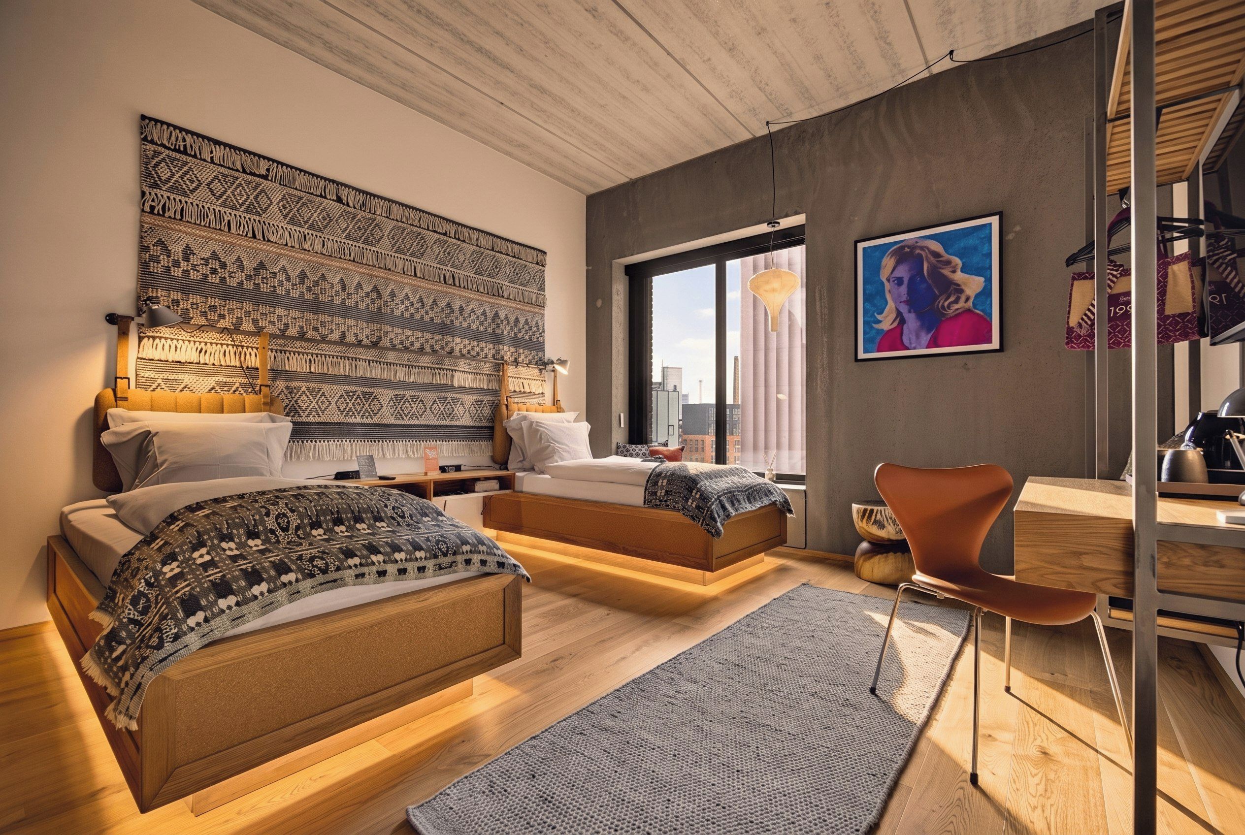 A modern bedroom with a large bed, a smaller bed, a desk, and a chair. There is a large window and a city view.