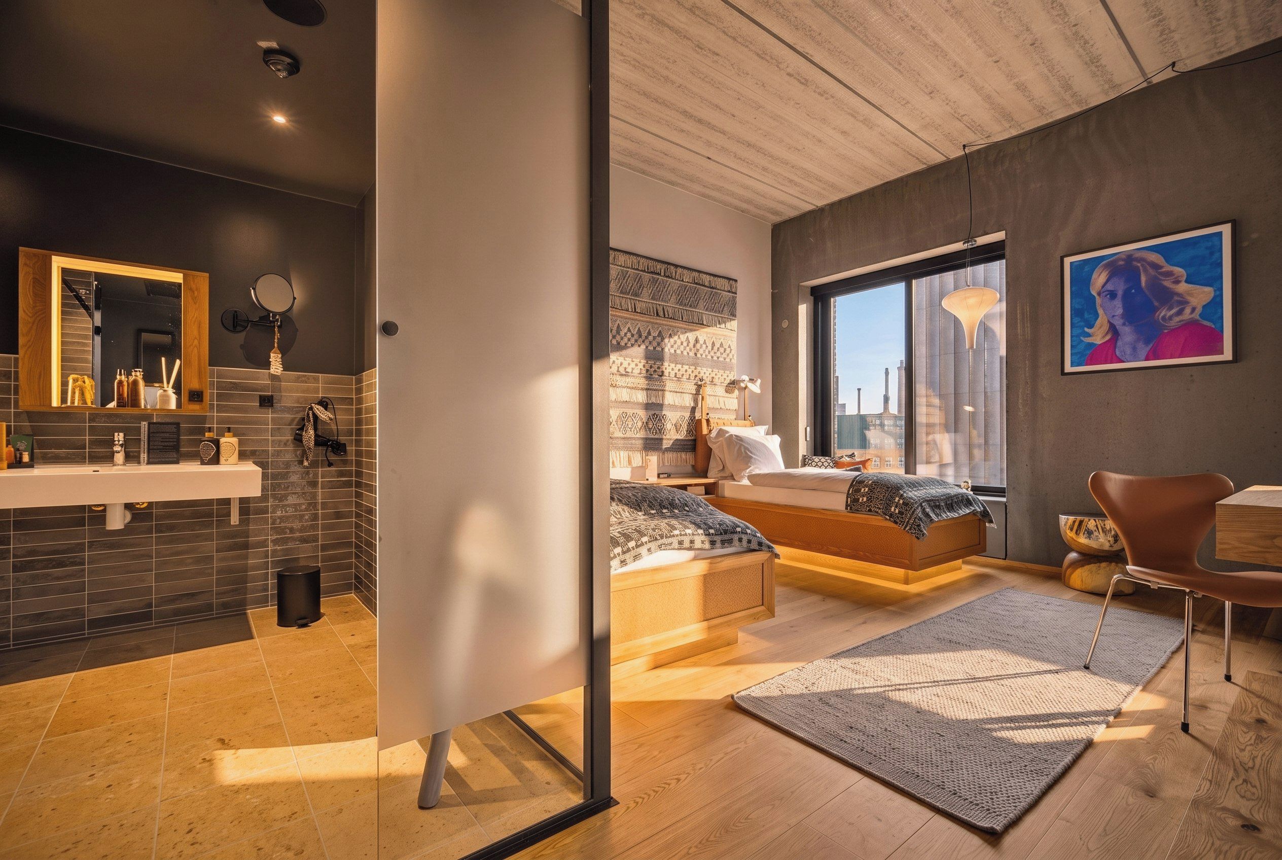 Un dormitorio moderno con una cama grande, vistas de la ciudad a través de una ventana y un baño con espejo y lavabo.
