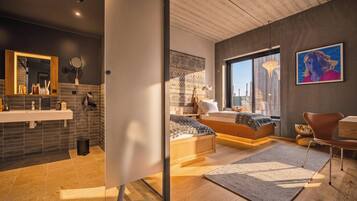 Un dormitorio moderno con una cama grande, vistas de la ciudad a través de una ventana y un baño con espejo y lavabo.
