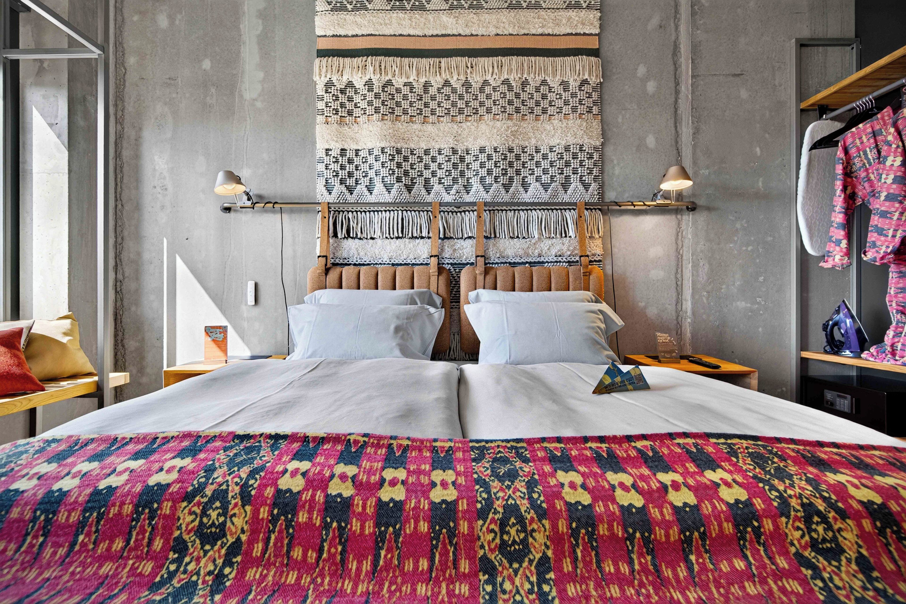 Un lit double avec une couvre-lit aux couleurs vives, une tête de lit ornée d’un motif tissé et deux lampes de chevet.