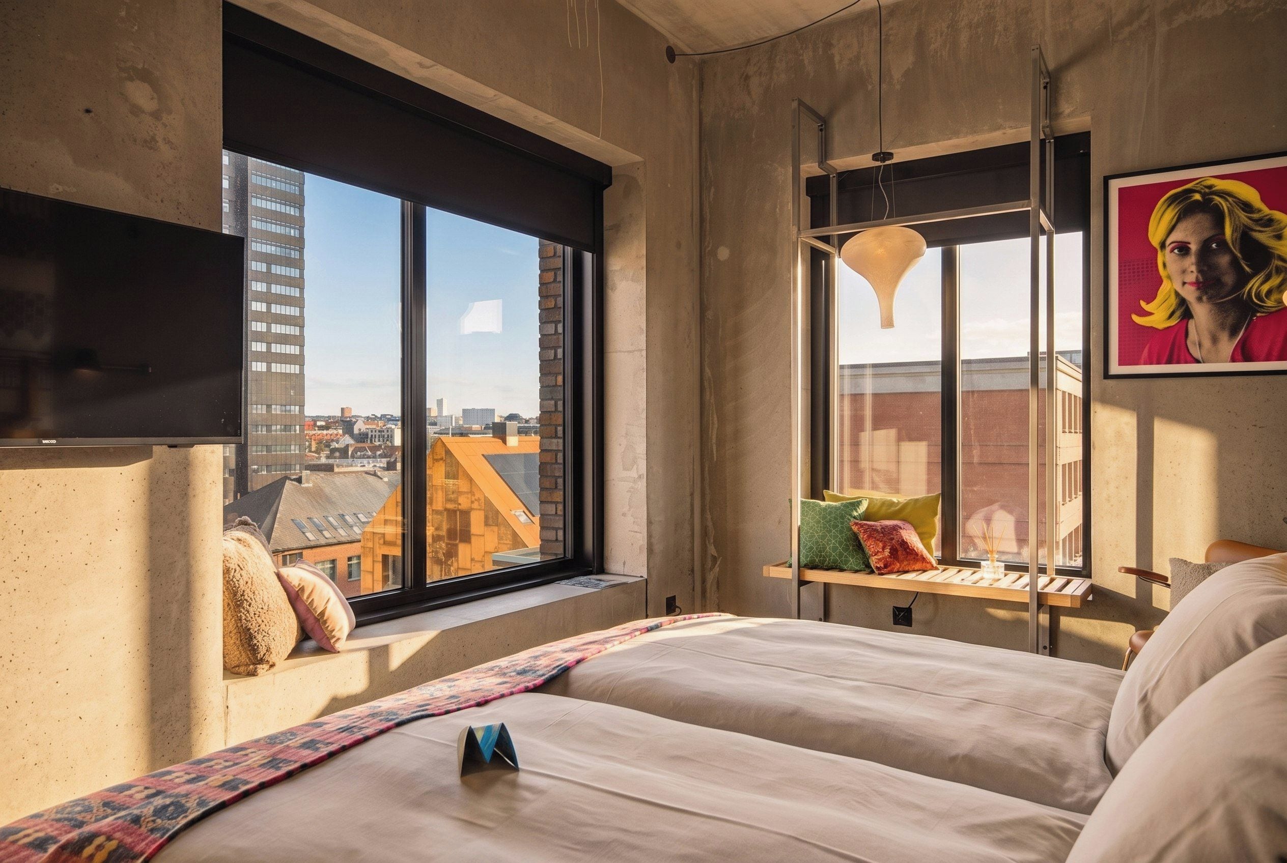 Un dormitorio moderno con un ventanal grande, una cama con almohadas, una lámpara colgante y vista a la ciudad.