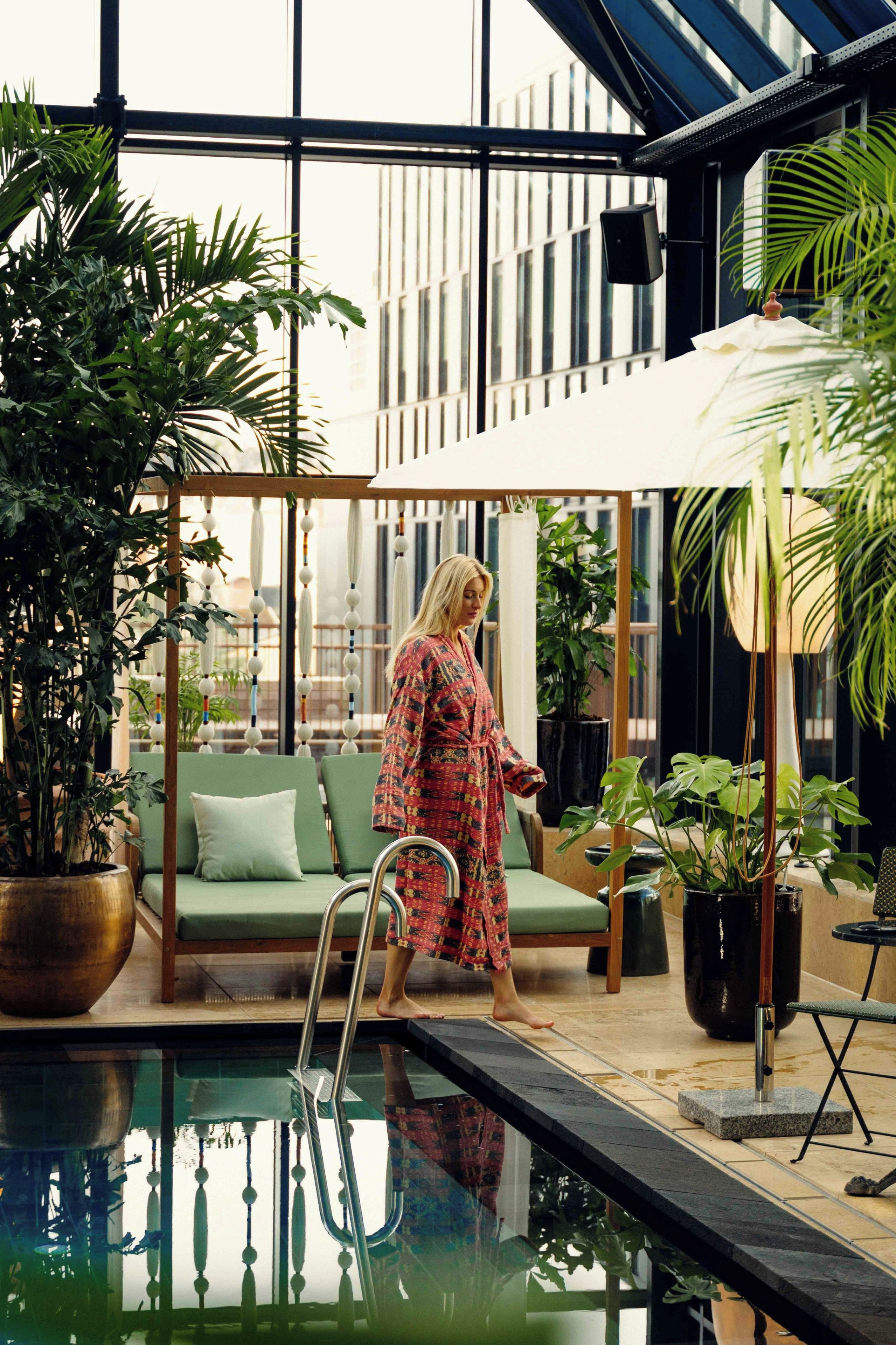 A woman walking by a pool with a glass ceiling, surrounded by indoor plants and a green sofa.