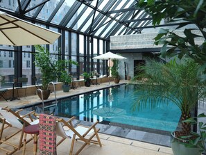 An indoor swimming pool with a glass ceiling, surrounded by potted plants and seating areas. - Belle Guldsmeden (Aarhus)