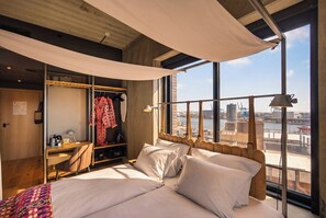 A hotel room with a large bed, a desk with a chair, a wardrobe, and a view of a cityscape through large windows. - Belle Guldsmeden (Aarhus)