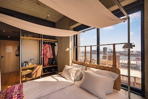 Habitación de hotel con una cama grande, un escritorio con silla, un armario y vistas a un paisaje urbano a través de amplias ventanas.