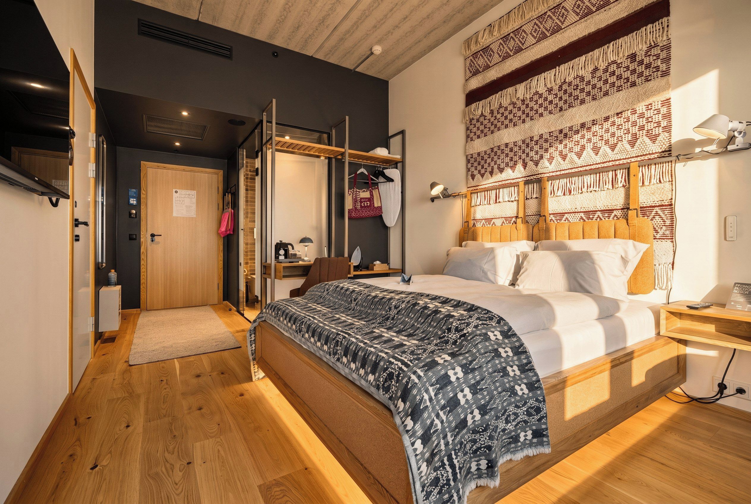 A modern bedroom with a large bed, a wooden floor, a wardrobe, a desk, and a patterned tapestry on the wall.
