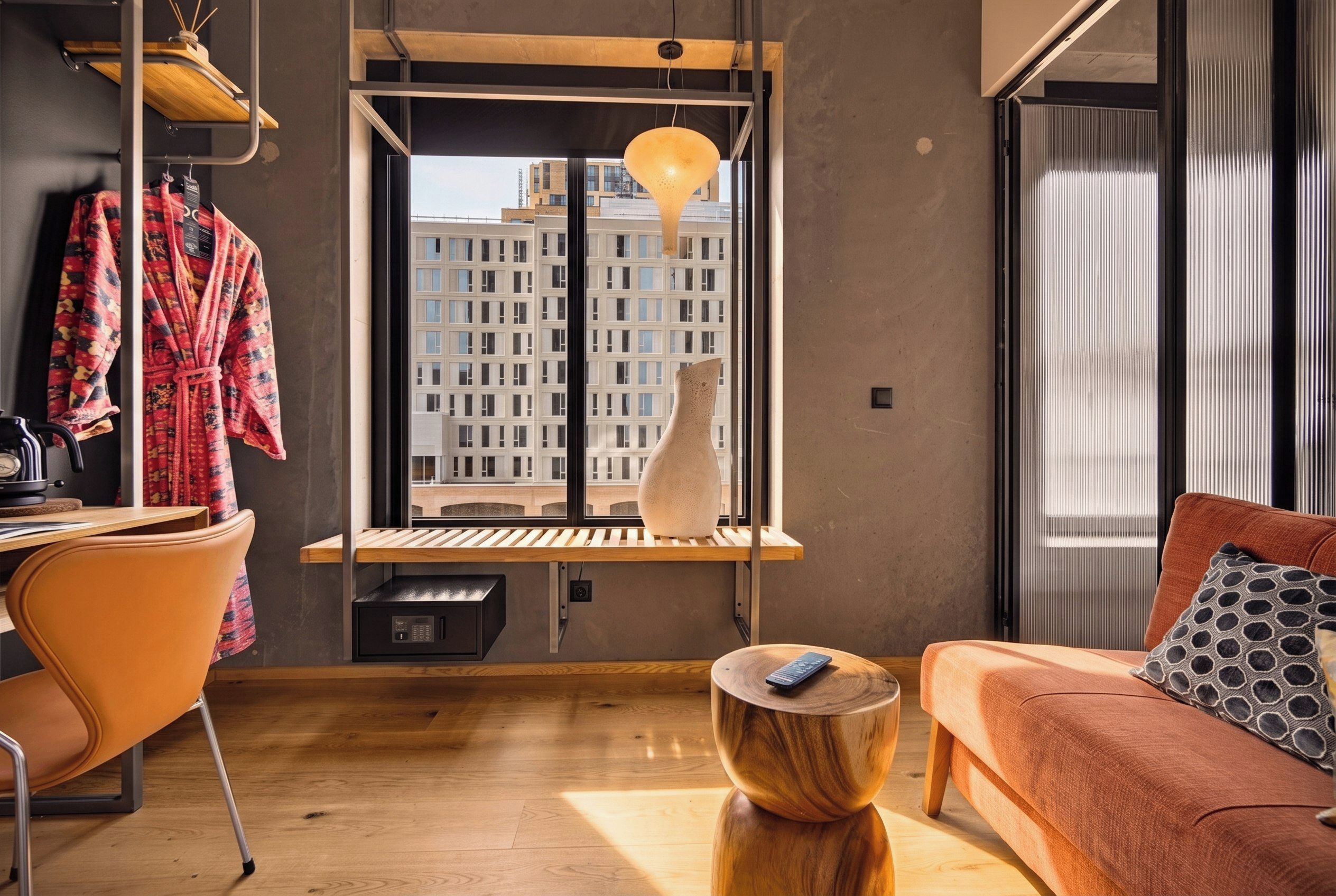 Uma sala de estar moderna com sofá, mesa de centro, poltrona, janela com vista para prédios e luminária pendente.