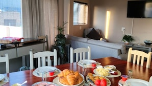 A dining area with a wooden table set for breakfast, white chairs, a TV mounted on the wall, and a window with blinds. - Moments Guesthouse (Kleinmond)
