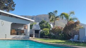 A pool with lounge chairs, a building with a patio, and a mountain in the background. - Moments Guesthouse (Kleinmond)