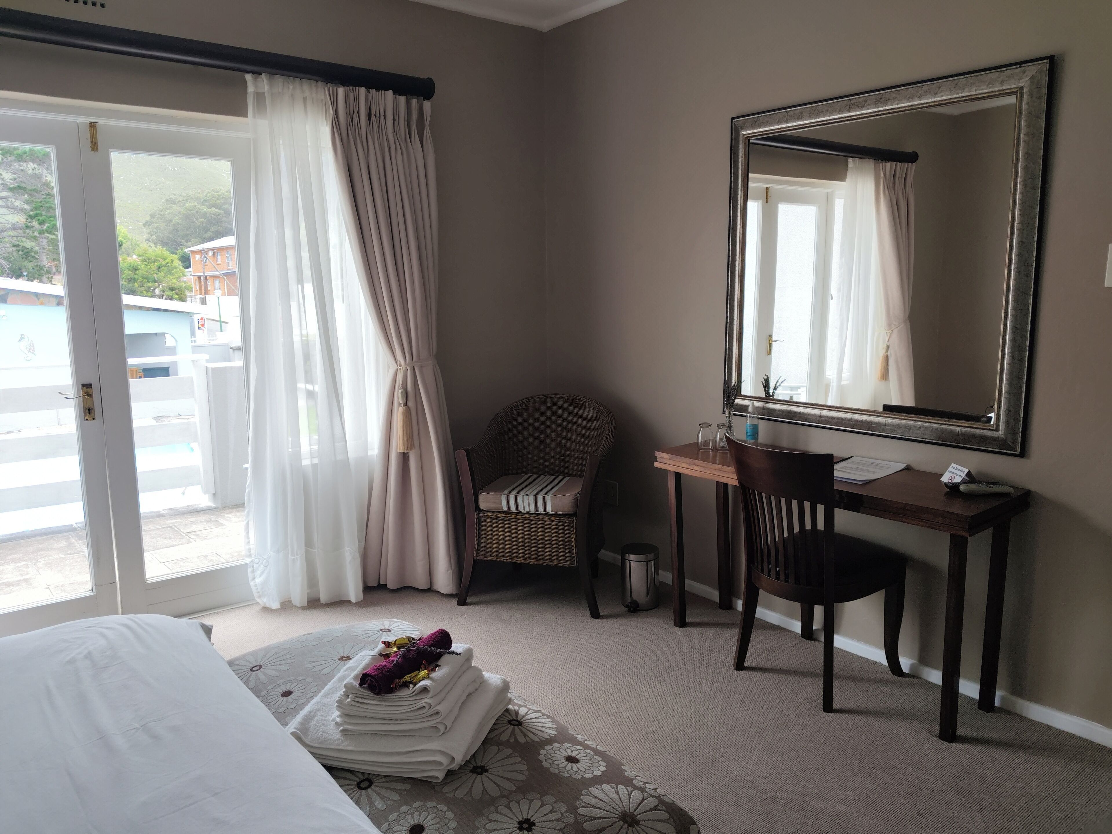 A hotel room with a bed, a desk with a chair, a large mirror, and a view through a window with curtains.