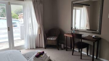 A hotel room with a bed, a desk with a chair, a large mirror, and a view through a window with curtains.