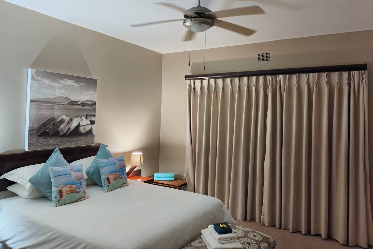 A hotel room with a bed, a ceiling fan, a nightstand with a lamp, a window with curtains, and a wall-mounted artwork.