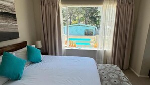 A bedroom with a large bed, a window with curtains, and a view of a pool and building outside. - Moments Guesthouse (Kleinmond)