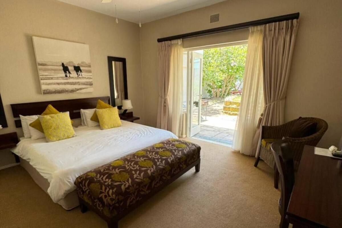 A hotel room with a bed, a bench, a chair, a desk, a window with curtains, and a painting on the wall.