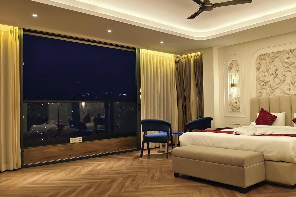 A hotel room with a large window, a bed with red pillows, a ceiling fan, and a wooden floor.