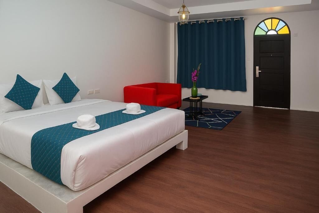 A hotel room with a bed, a red armchair, a small table with a vase, and a door with a stained glass window.