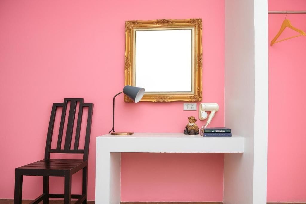 Une pièce avec un mur rose, une chaise en bois, un bureau blanc, une lampe, un miroir encadré et un cintre.