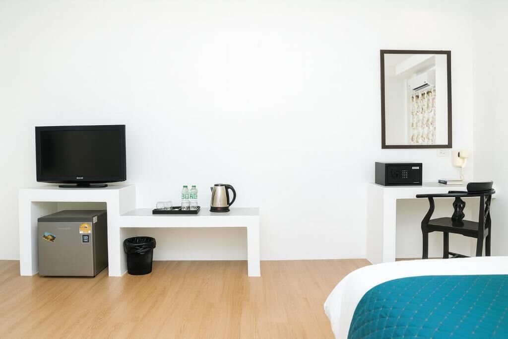 A hotel room with a bed, a television, a mini-fridge, a coffee maker, a trash bin, a small table with a chair, and a framed picture on the wall.