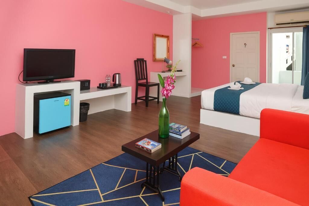 A modern hotel room with a pink wall, a flat-screen TV, a desk with a computer, a coffee machine, a chair, a bed with a blue blanket, and a red sofa.