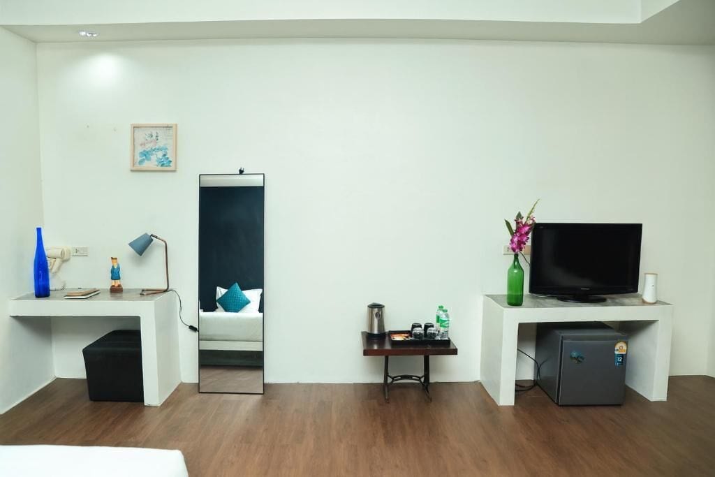 A hotel room with a white desk, a television, a small refrigerator, and a bed with a blue pillow.