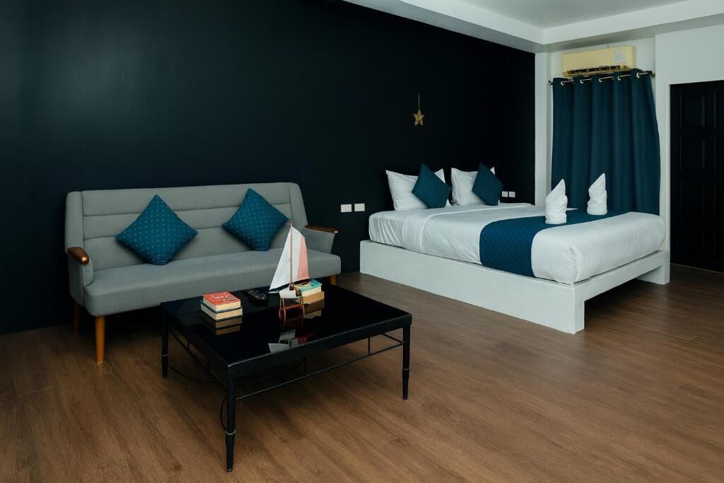 Una habitación de hotel moderna con un sofá gris, una cama con ropa de cama blanca, una mesita de centro negra con libros y una lámpara, y una pared oscura con decoración de estrellas.