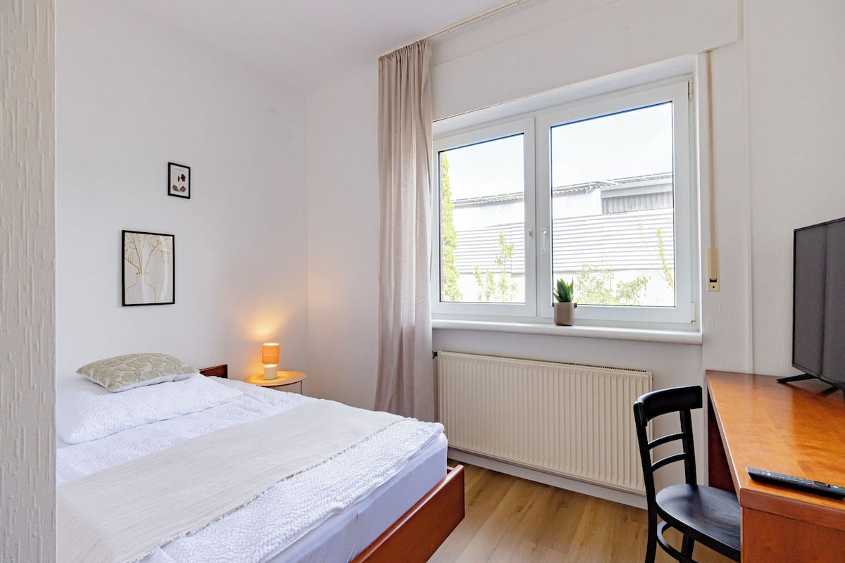 Un dormitorio con cama, escritorio, silla, televisor, ventana con cortinas y radiador.