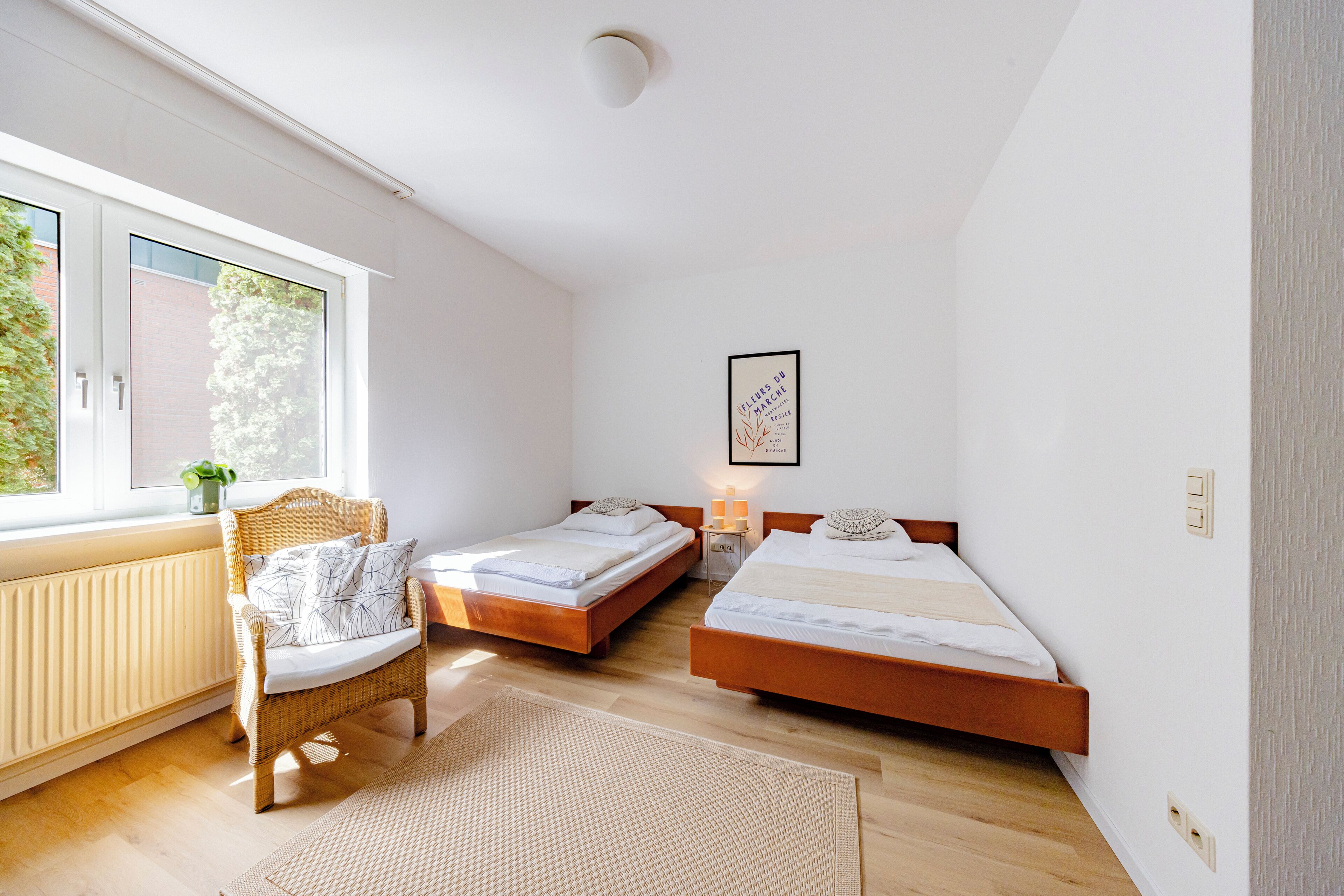 A bedroom with two beds, a wicker chair, a radiator, a window, and a framed picture on the wall.