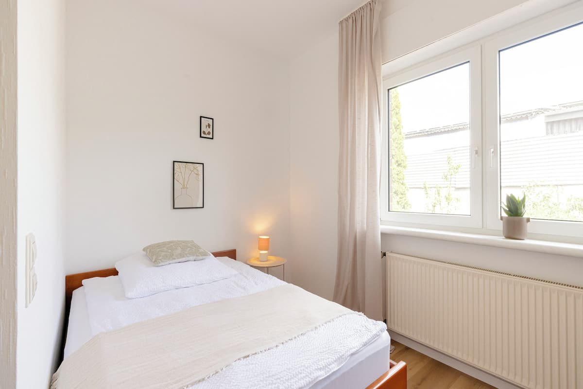 Un dormitorio con cama, lámpara de noche, ventana con cortinas y radiador.