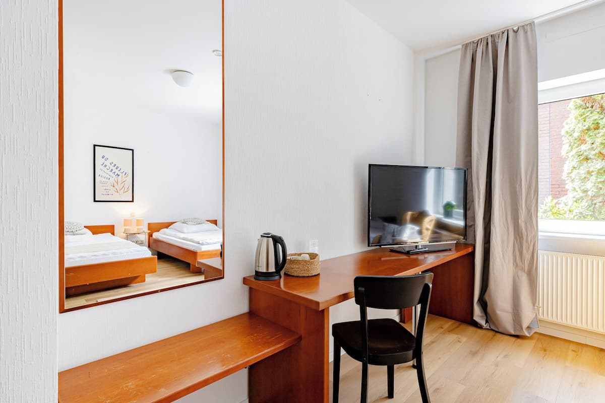 Habitación de hotel con cama, escritorio con televisor, silla, ventana con cortinas y radiador.