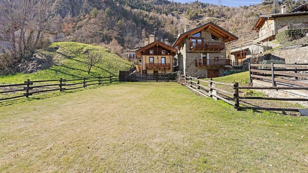 Un bâtiment de style chalet, doté d’un balcon en bois et entouré d’une clôture en bois, se dressant sur un fond de paysage montagneux.