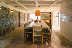 A long dining table with chairs, stone walls, and a hanging light fixture. - Villa Estasia front sea - My Rental Homes (Begur)