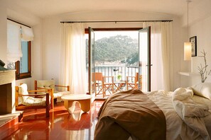 A hotel room with a bed, a seating area with chairs, a small table, and a balcony with a view of a mountainous landscape. - Villa Estasia front sea - My Rental Homes (Begur)