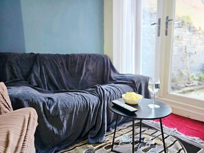A cozy living area with a dark-colored sofa, a small round table with a bowl of snacks and a glass of water, and a remote control. - 3-Bedroom Town Centre House | Sleeps 8 | Self Check-in | Parking | Pet Friendly (St Helens)
