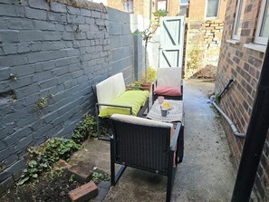 En smal udendørs plads med siddeområde bestående af en fletstol og et lille bord med kop og underkop.