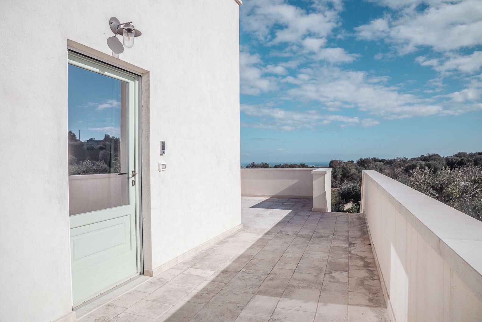 A balcony with a door, a light fixture, and a view of the sky and vegetation.
