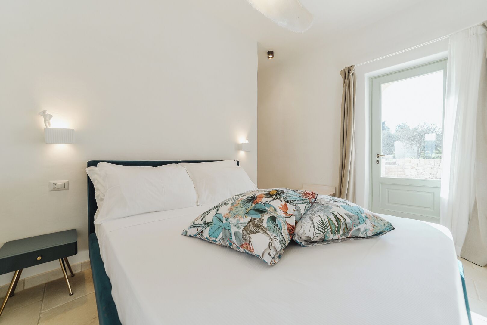 A bedroom with a bed, a nightstand, a wall-mounted light, and a window with curtains.