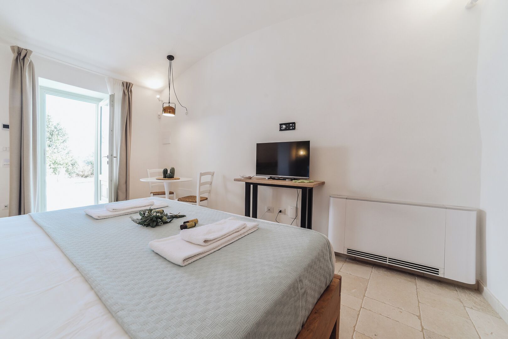 Habitación de hotel con cama, televisor en un mueble bajo, una mesita con un cactus y ventana con cortinas.