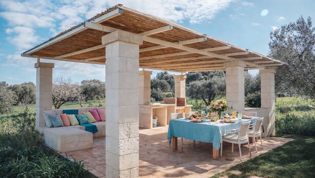 An outdoor patio with a wooden pergola, stone pillars, a table set for dining, and a cushioned seating area. - DIMORA STUNE (Ostuni)