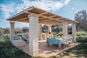 An outdoor patio with a wooden pergola, stone pillars, a table set for dining, and a cushioned seating area. - DIMORA STUNE (Ostuni)