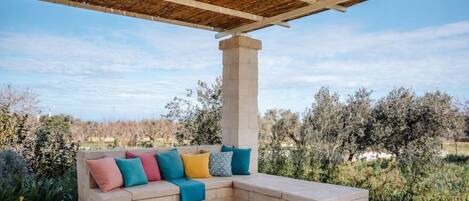 A covered outdoor seating area with a stone bench and colorful cushions, surrounded by vegetation and a clear sky.