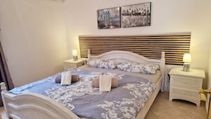 A neatly made bed with a patterned bedspread, two pillows, and rolled towels. There are two bedside tables with lamps, and a wall-mounted air conditioning unit. - Charming finca with pool and large garden in an idyllic setting in Xaló (Teulada)