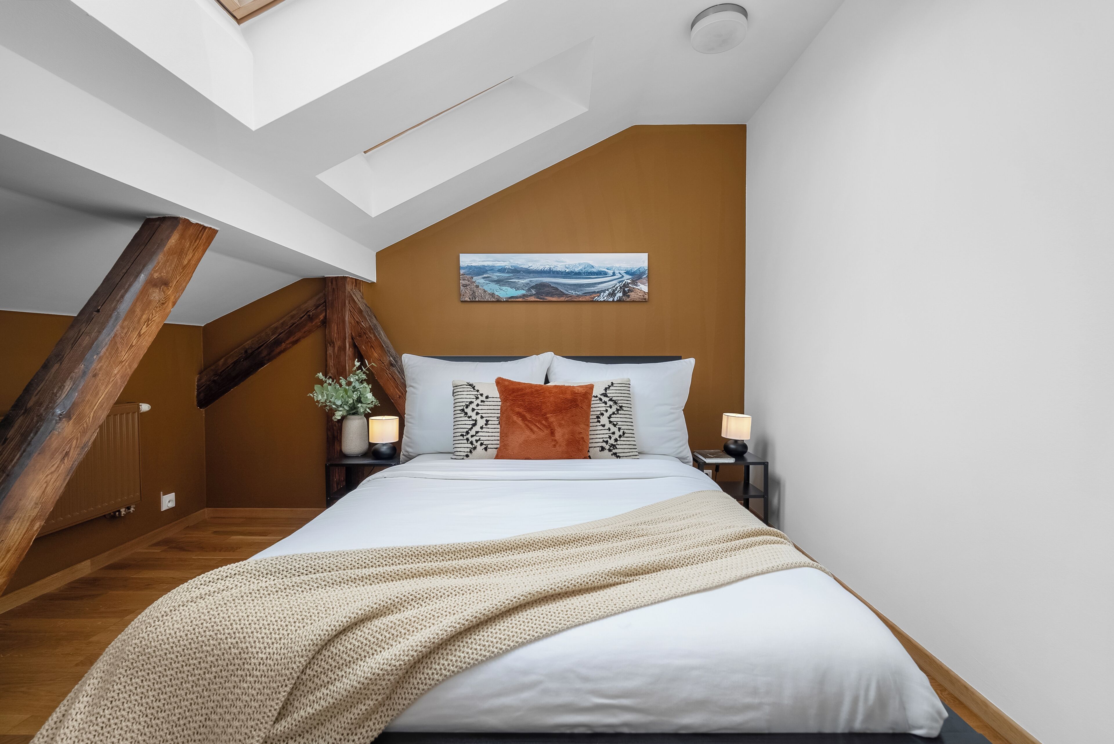 A bedroom with a sloped ceiling, a bed with white bedding, a brown wall, and a painting of a landscape.