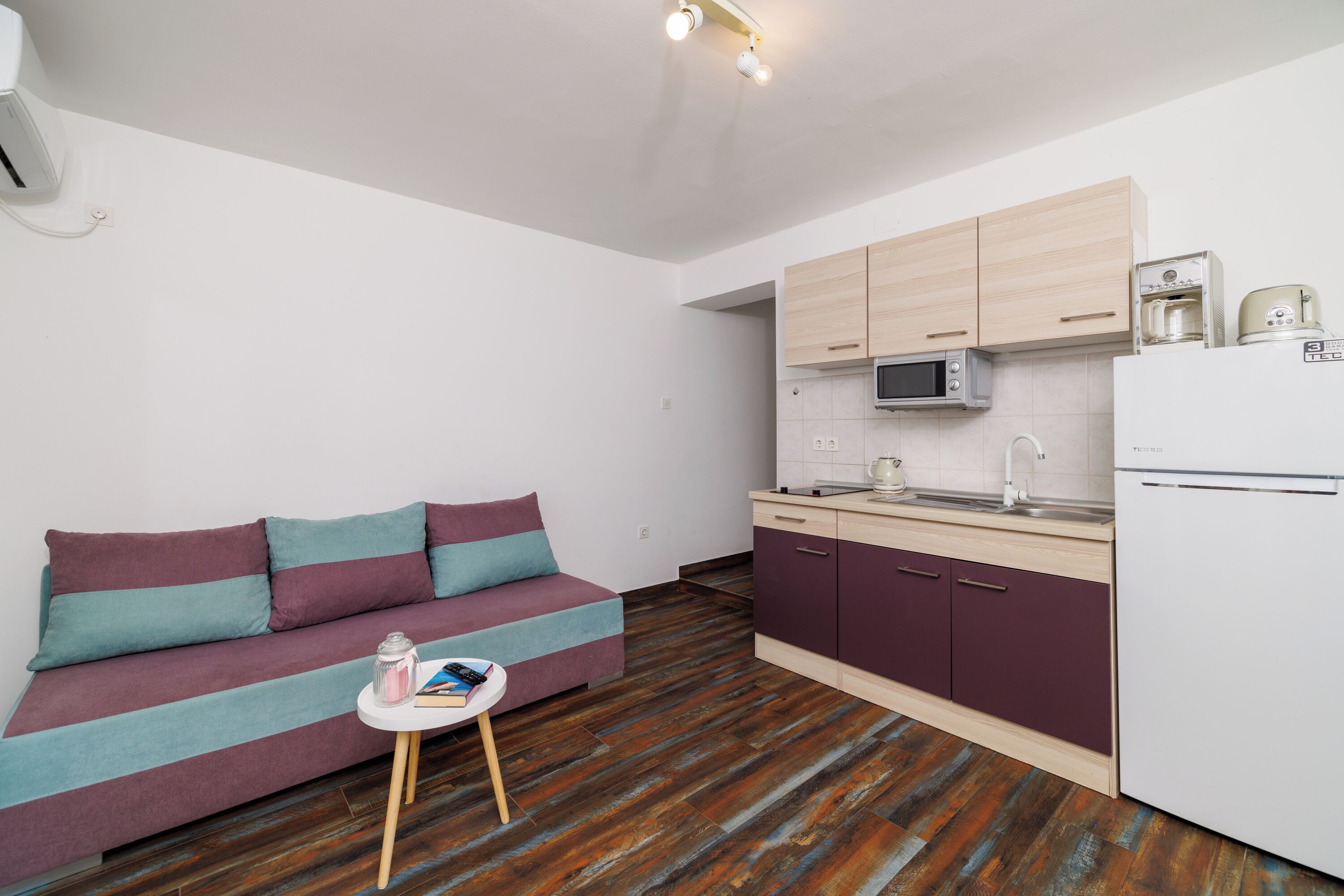 Cocina compacta con microondas, tostadora y refrigerador. En la sala de estar, un sofá con cojines a rayas y una pequeña mesa con un libro y una vela.