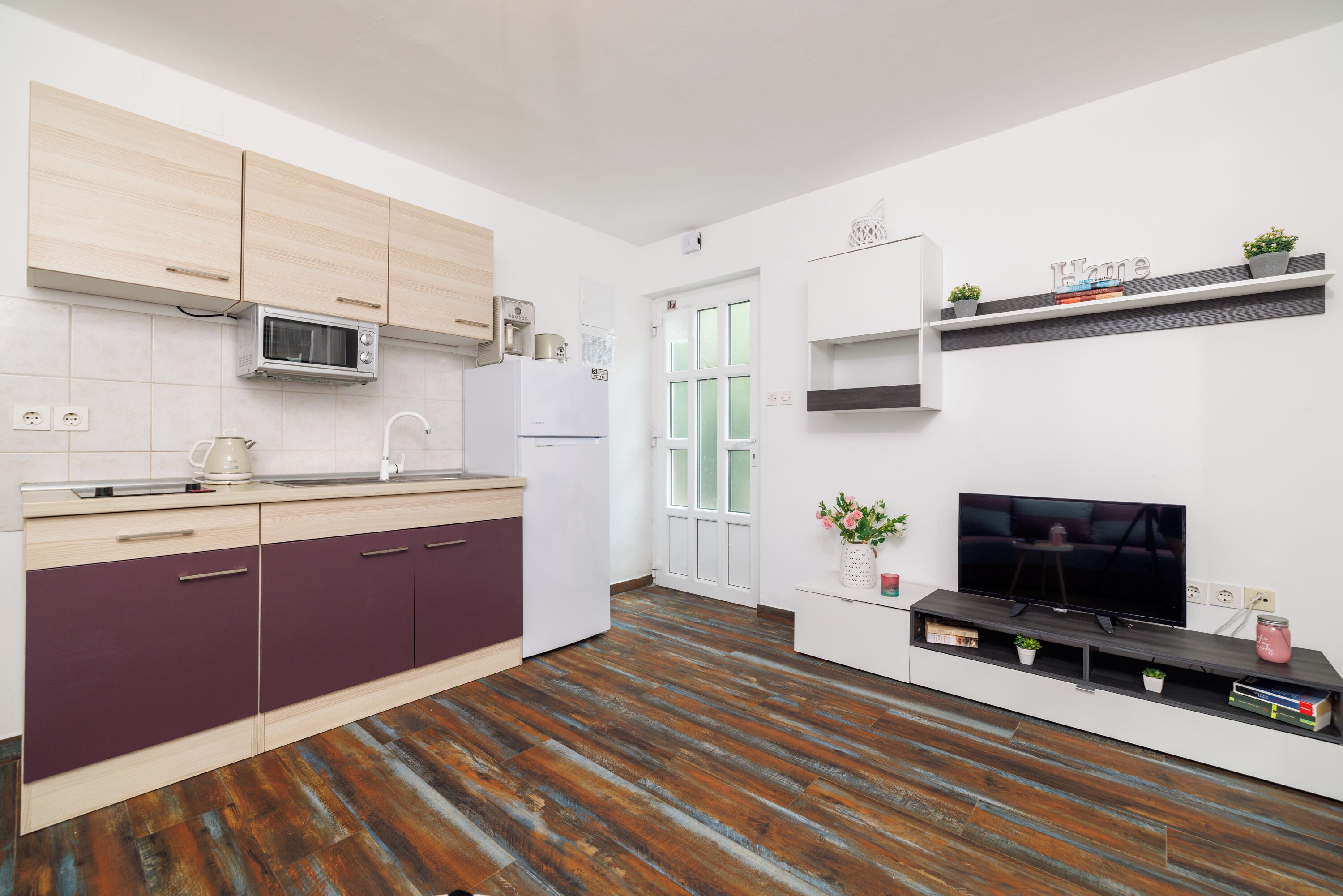 A modern kitchen with wooden cabinets, a microwave, and a refrigerator. There is a TV on a stand and a shelf with plants and books.