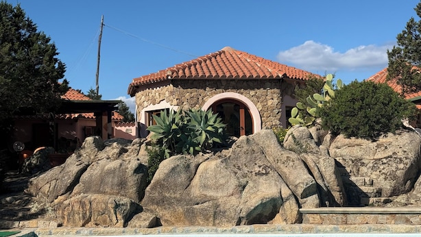 A stone building with a red-tiled roof, a pool, and large rocks in the foreground.