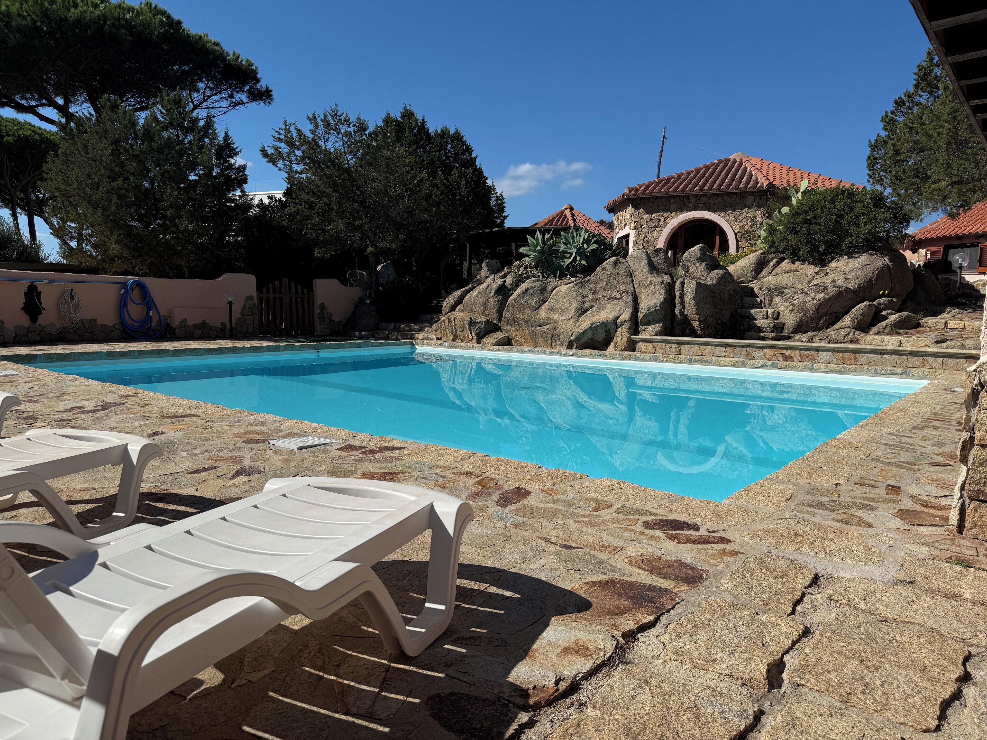 A swimming pool with stone tiles, lounge chairs, and a stone building with a rounded archway.